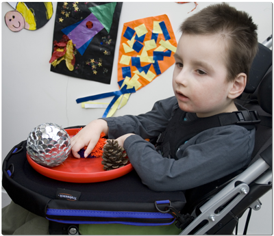 Image of a young boy in a adapted buggy, playing with sensory items on a blue trim Trabasack Curve Connect wheelchair tray and bag