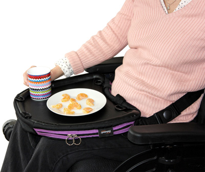 Image of a lady wearing a salmon-coloured jumper, sat in a wheelchair with a purple-trim Trabasack Curve tray bag on her knee, with a cup and plate of tangerine on top